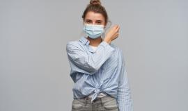 teenage-girl-happy-looking-woman-with-blond-hair-gathered-in-bun-wearing-striped-knotted-shirt-and-protective-face-mask-takes-mask-off-looking-at-the-camera-isolated-over-grey-wall.jpg