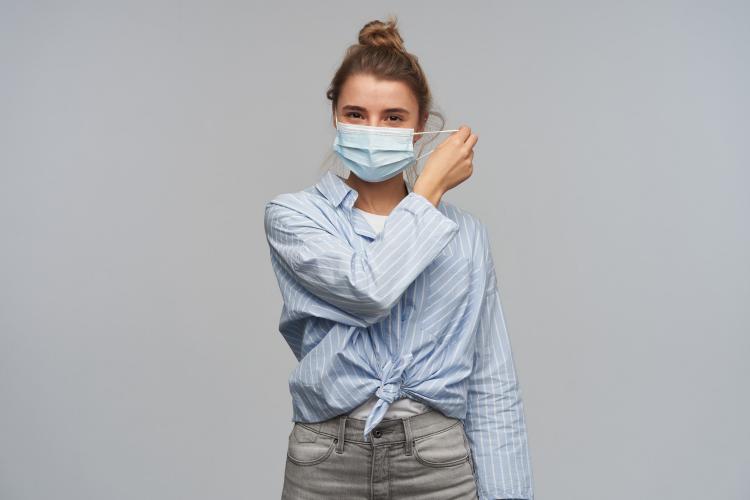 teenage-girl-happy-looking-woman-with-blond-hair-gathered-in-bun-wearing-striped-knotted-shirt-and-protective-face-mask-takes-mask-off-looking-at-the-camera-isolated-over-grey-wall.jpg