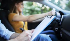 close-up-view-of-driving-instructor-holding-checklist-while-in-background-female-student-steering-and-driving-car.jpg