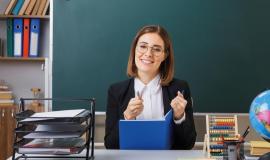 young-woman-teacher-wearing-glasses-sitting-school-desk-front-blackboard-classroom-checking-class-register-looking-camera-smiling-cheerfully_141793-132944.jpg