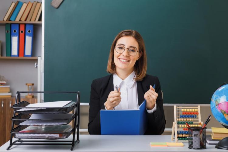 young-woman-teacher-wearing-glasses-sitting-school-desk-front-blackboard-classroom-checking-class-register-looking-camera-smiling-cheerfully_141793-132944.jpg